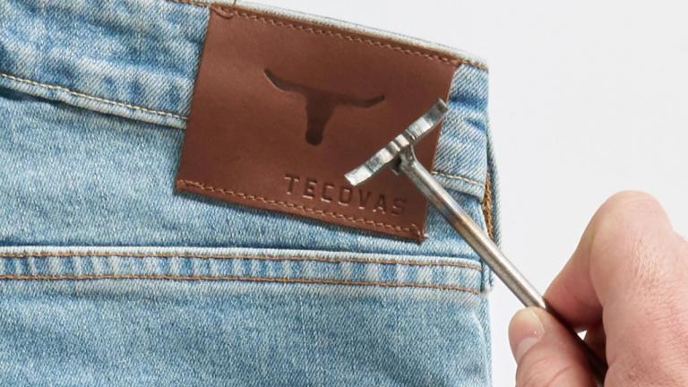 A branding iron is about to press onto the brown leather patch labeled "Tecovas" on the waistband of light blue denim jeans.