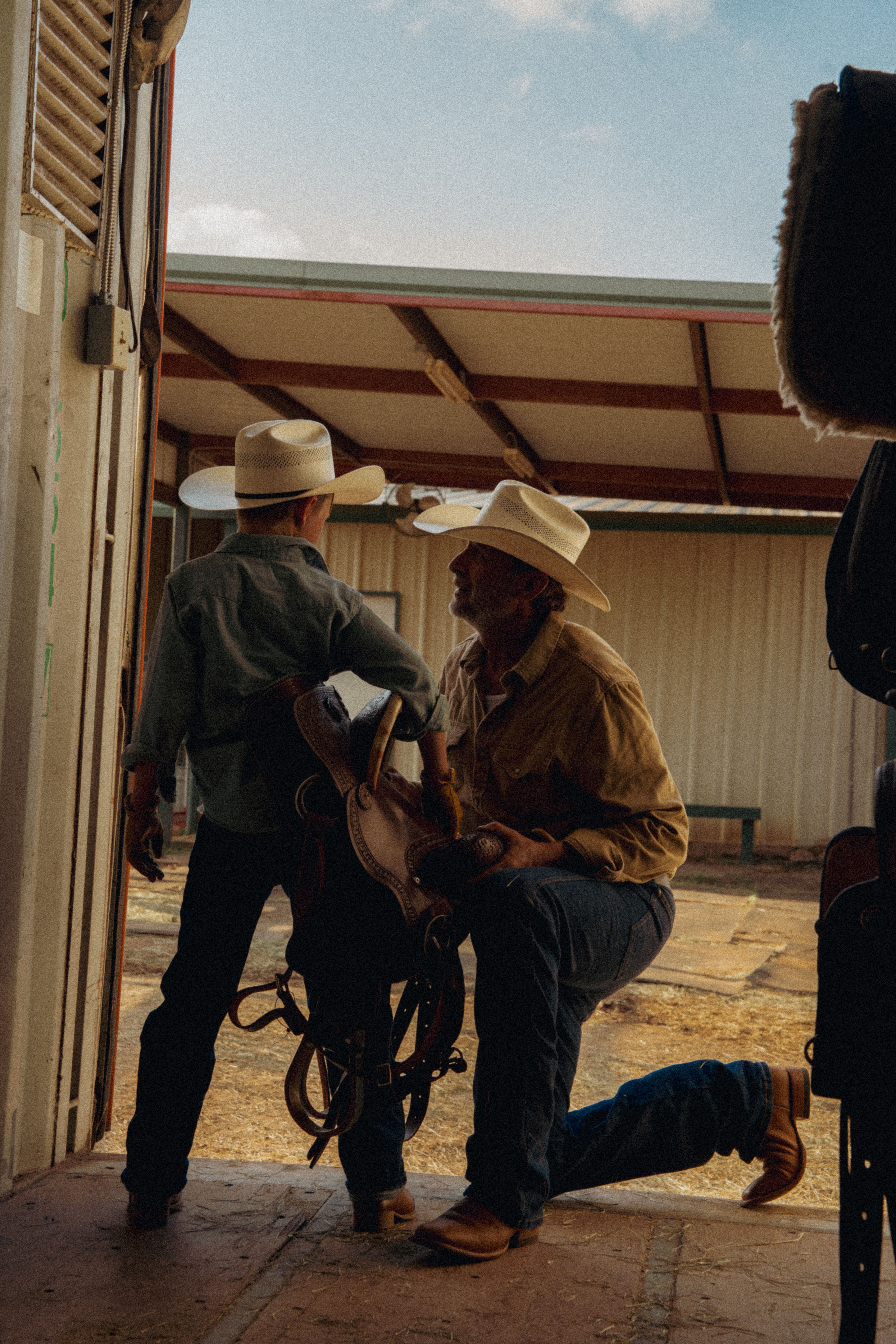 An adult and a child wearing cowboy hats stand in a barn doorway, with the adult kneeling and holding a saddle while the child looks outside.