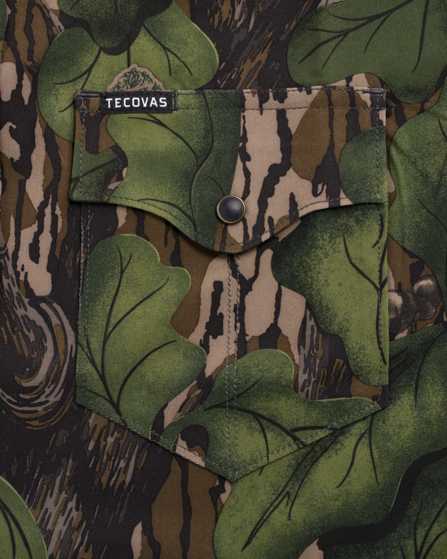 Close-up of a shirt pocket with a green leaf and brown tree bark camouflage pattern, featuring a black snap button and a Tecovas brand label.