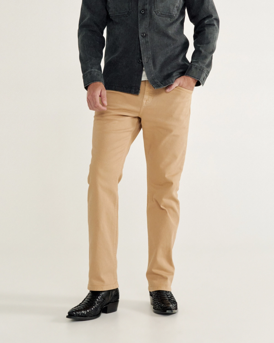 A person in a black button-up shirt and tan pants stands with one hand in a pocket. They wear black textured shoes.