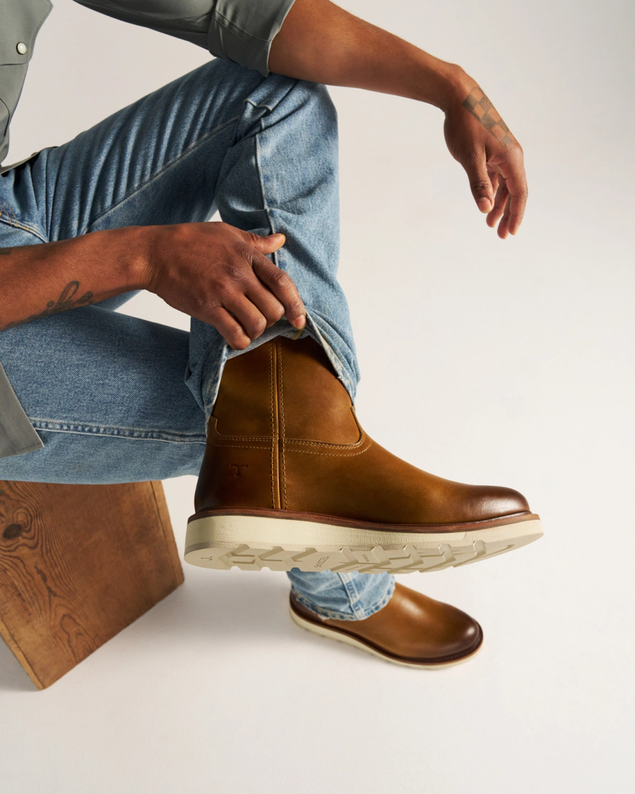 The Knox in Wheat Cowhide. A person in jeans and a gray shirt sits on a wooden bench, crossing their leg and wearing wheat cowhide brown leather boots with thick white soles.