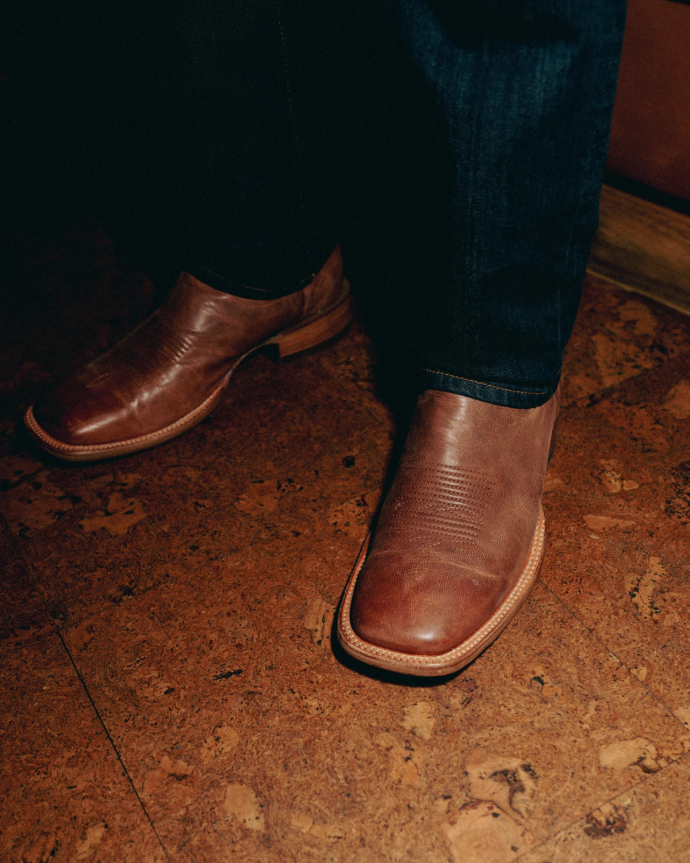 Person wearing brown leather shoes and dark jeans, standing on a wooden floor.