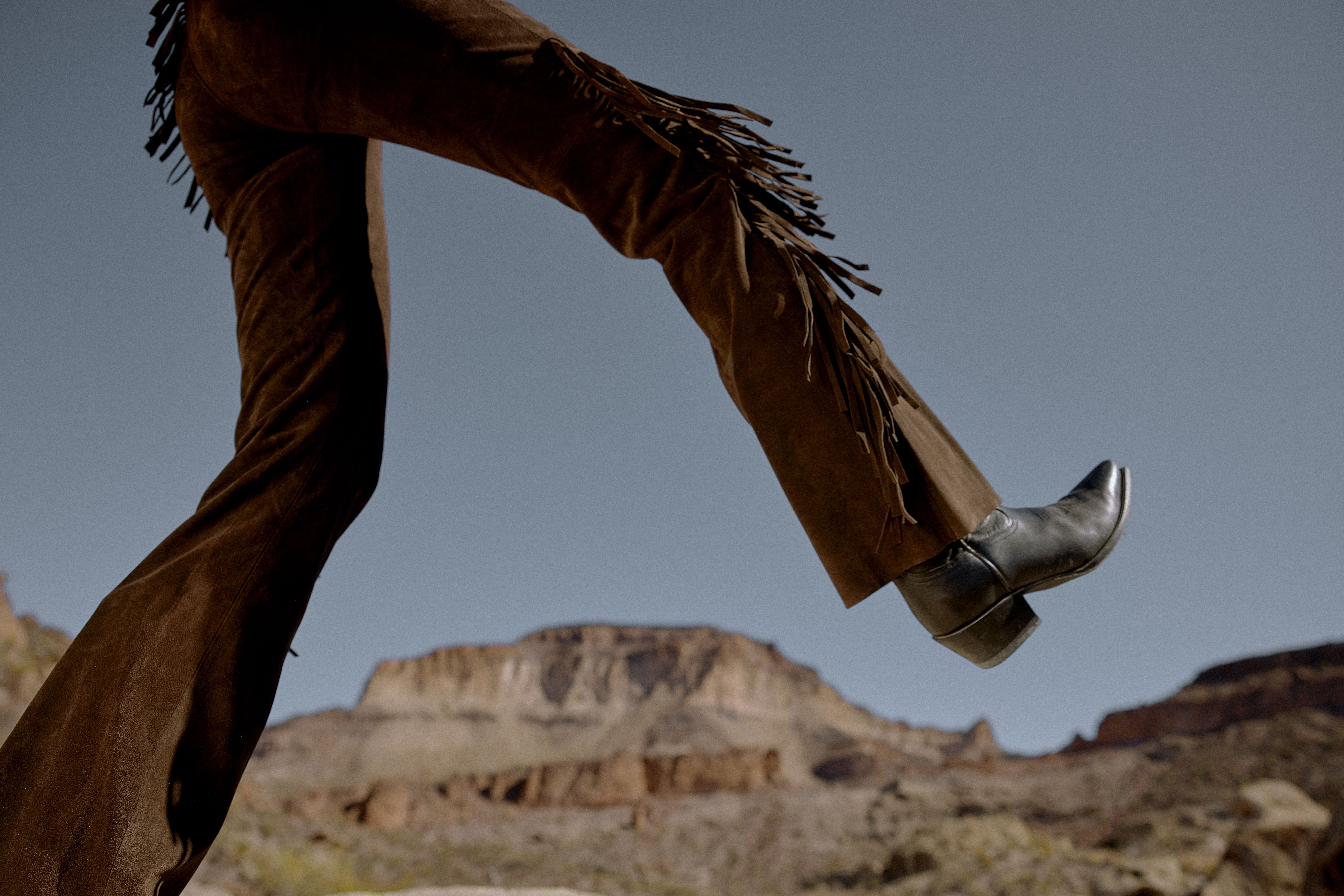 Person wearing brown fringe pants and black heeled boots steps forward with a desert landscape and rocky cliffs in the background under a clear sky.