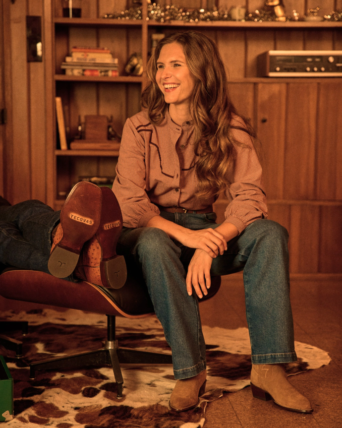 A woman with long hair sits smiling in a wooden-paneled room, wearing jeans and Caramel Suede Tecovas boots—The Jamie Suede style—with one leg propped up on a chair.