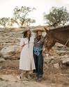 Two people in Western attire stand beside a saddled horse on a rocky terrain with a dog nearby. Trees and a setting sun are in the background.