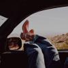 Person wearing blue jeans and red cowboy boots rests their feet outside a car window, with a desert landscape visible in the background.