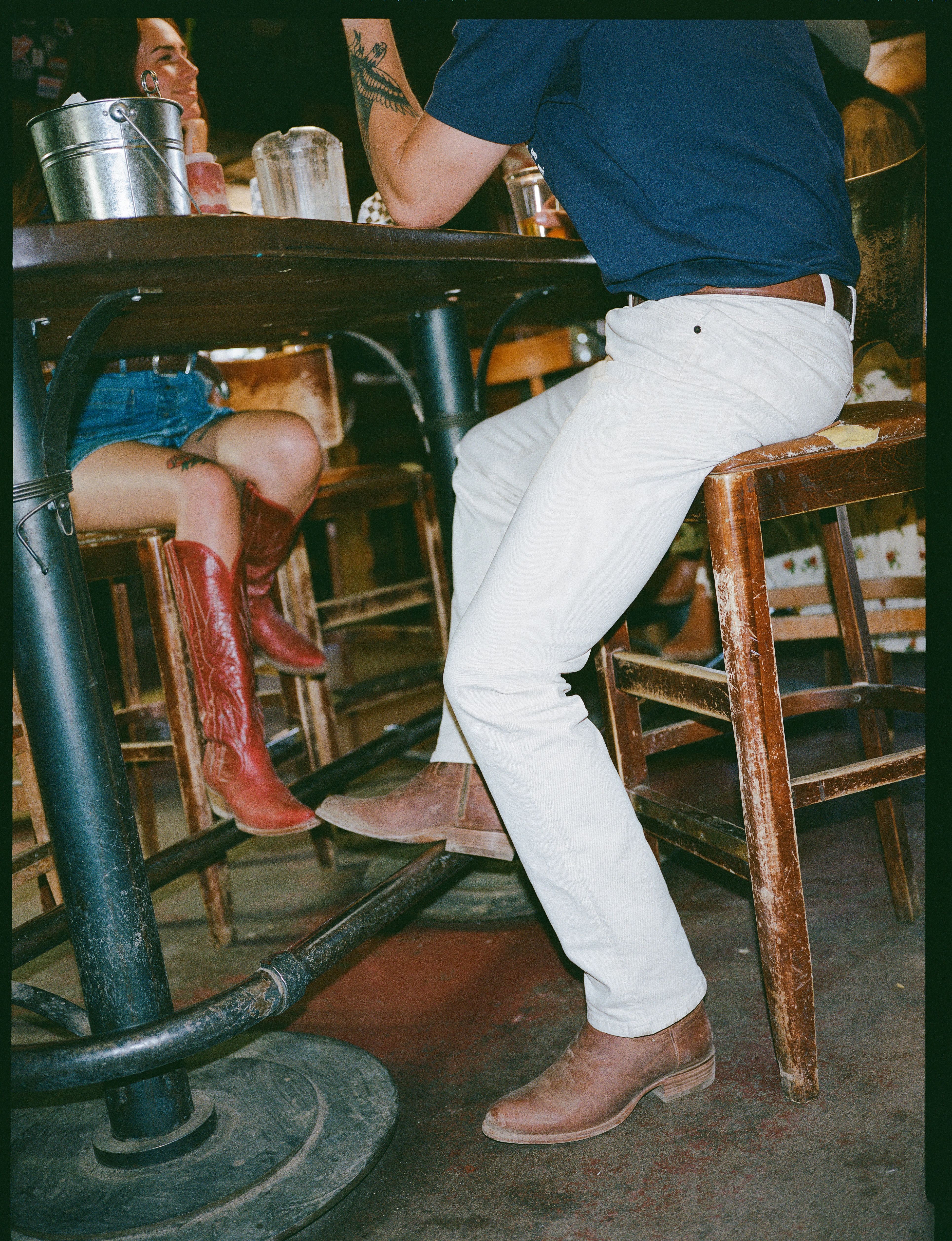 Two people sit at a wooden table in a casual setting; one wears red cowboy boots and denim shorts, the other white pants, brown boots, and a navy shirt. Only their lower bodies are visible.