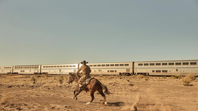 A person wearing a cowboy hat rides a horse across a dry, dusty landscape as a long passenger train passes by in the background.