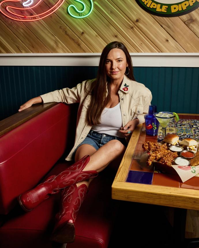 A woman in a white top, denim shorts, and red cowboy boots sits in a restaurant booth with food and drinks on the table.