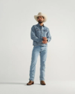 A man wearing a white cowboy hat, denim jacket, denim jeans, and brown cowboy boots stands against a plain light background.