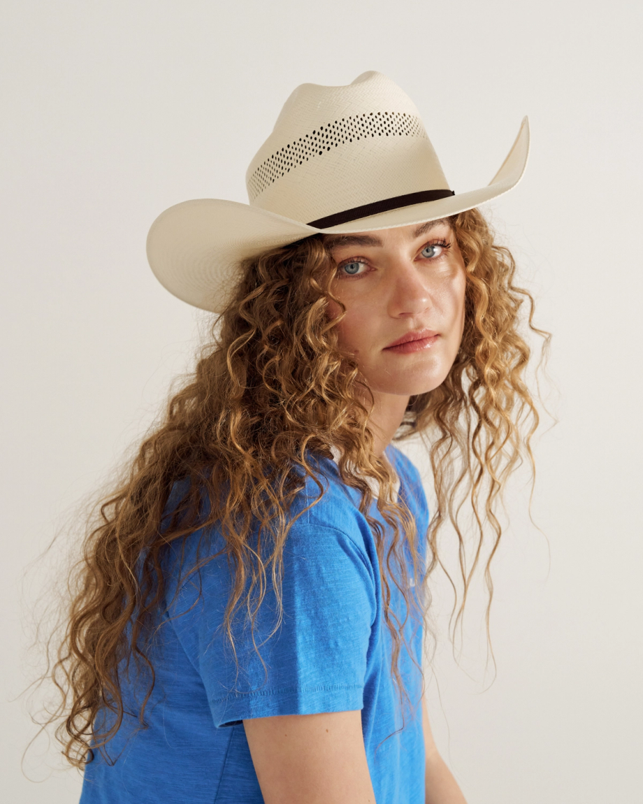Woman with long curly hair wearing a blue T-shirt and a light-colored cowboy hat, looking toward the camera against a plain background.