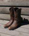 A pair of worn brown leather cowboy boots is placed side by side on a wooden porch in sunlight, casting distinct shadows.