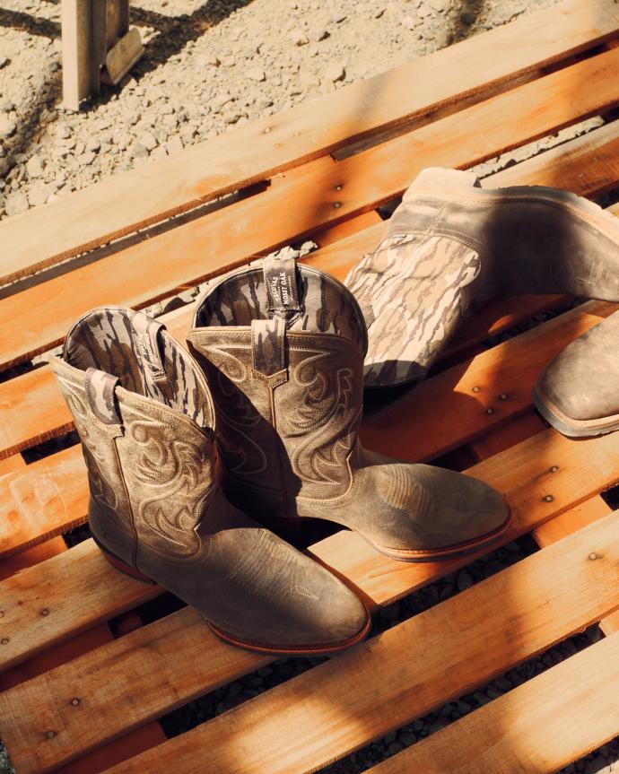 A pair of brown cowboy boots with decorative stitching sit on a wooden pallet in sunlight.