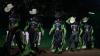 Five rodeo competitors wearing matching black and green outfits and cowboy hats walk in a dimly lit arena.