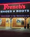 Storefront of French’s Shoes & Boots with illuminated signage above wooden exterior, displaying window merchandise and entrance doors at night.