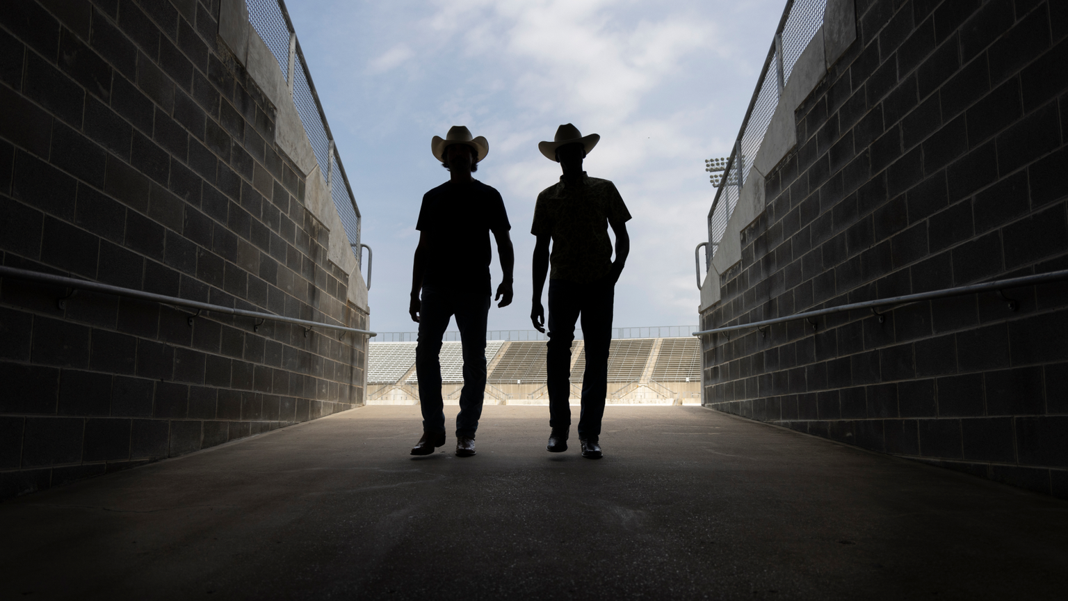 Two silhouetted figures wearing cowboy hats walk down a pathway between brick walls, leading towards an open stadium with a partly cloudy sky overhead.