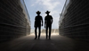 Two silhouetted figures wearing cowboy hats walk down a pathway between brick walls, leading towards an open stadium with a partly cloudy sky overhead.