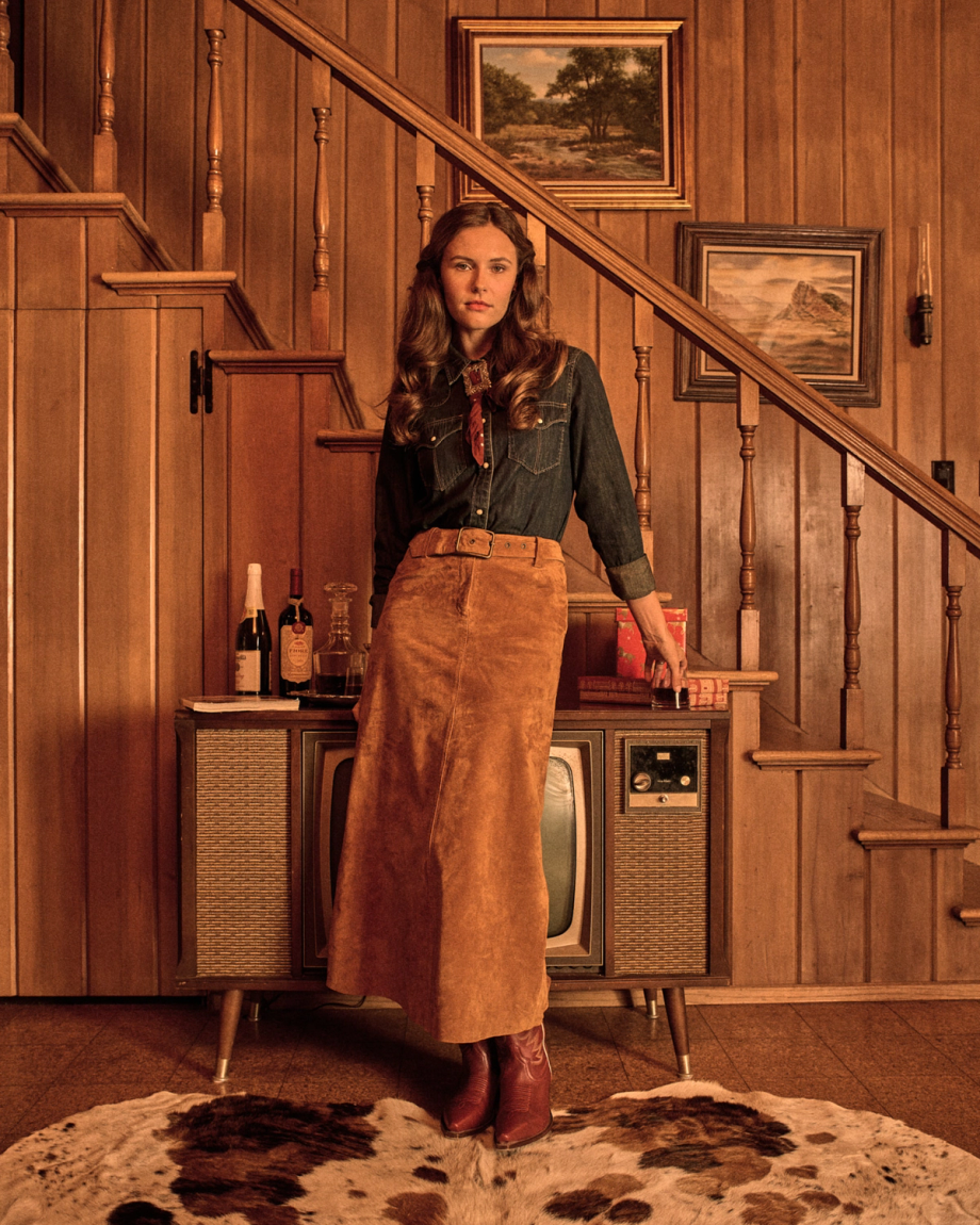 A woman in a denim shirt, brown skirt, and boots stands indoors by a vintage TV, in a wood-paneled room with drinks, art, and a cowhide rug.