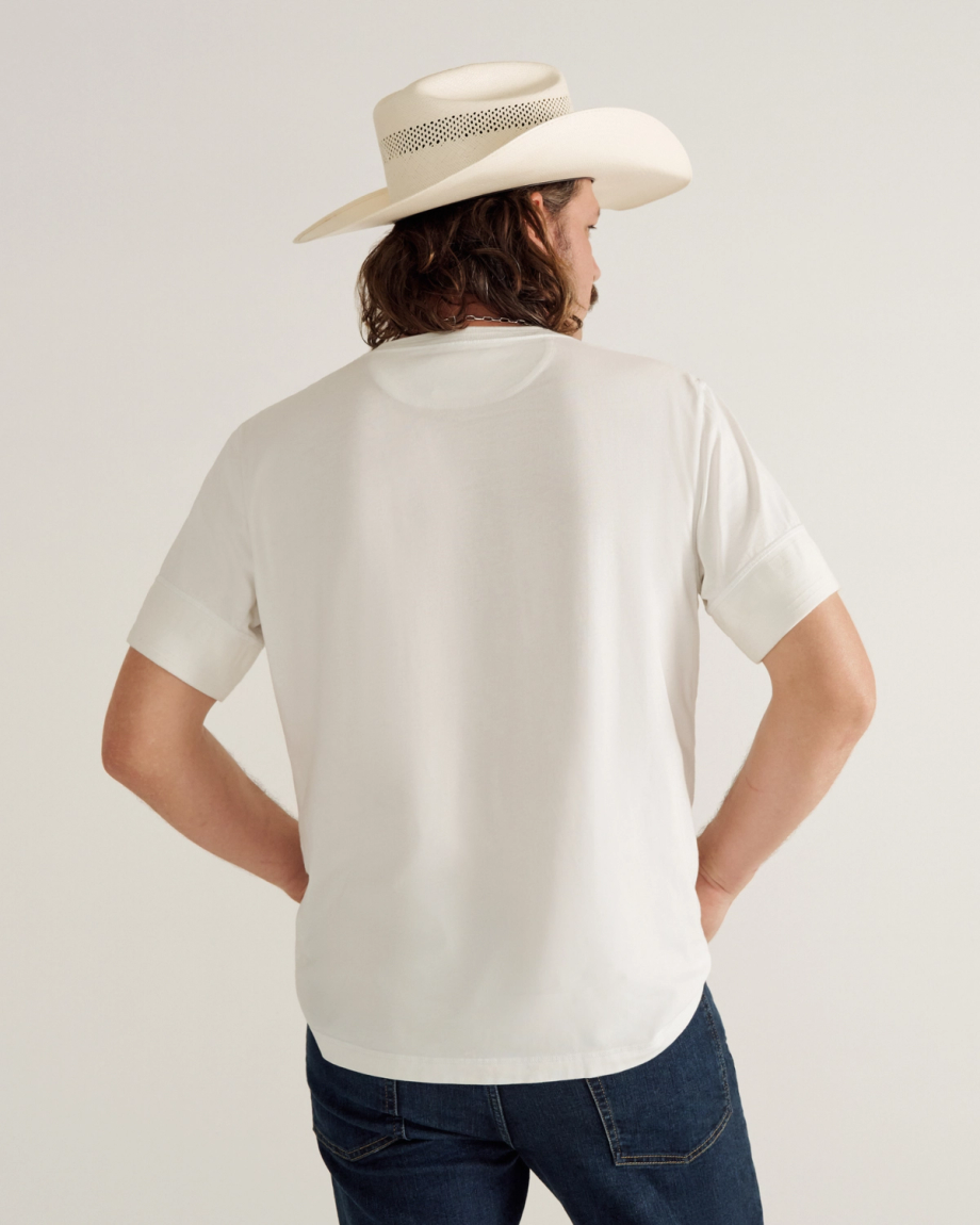 A person wearing a white short-sleeve t-shirt, blue jeans, and a cream-colored cowboy hat stands facing away from the camera against a plain background.