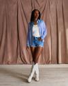 A woman stands in front of a brown backdrop, wearing a blue plaid shirt, white tank top, denim shorts, and white cowboy boots.