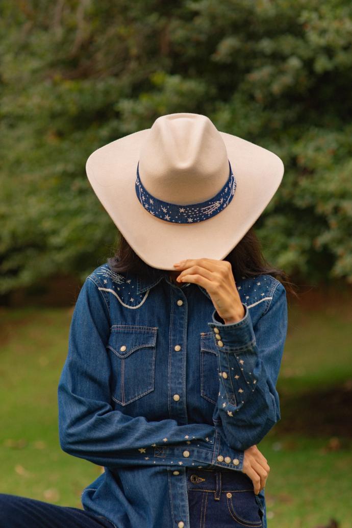 Person wearing a wide-brimmed hat with a blue pattern band and a denim shirt, covering their face with the hat brim, standing outdoors with greenery in the background.