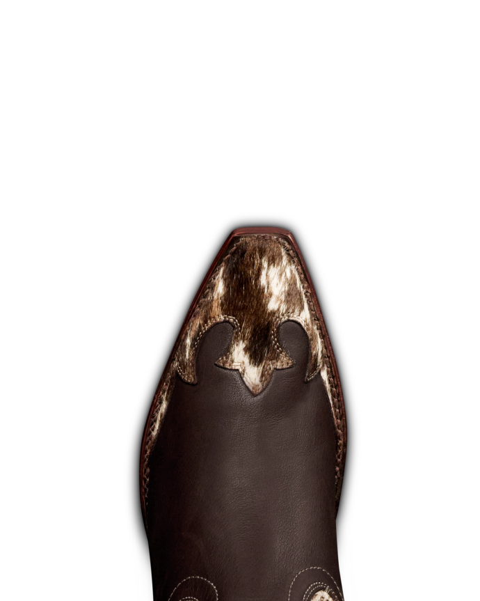 The Sadie in Hair on Hide Hair on Hide / Cowhide. Close-up of the pointed toe of a brown leather cowboy boot with decorative stitching and a patch of textured, spotted Hair on Hide cowhide.