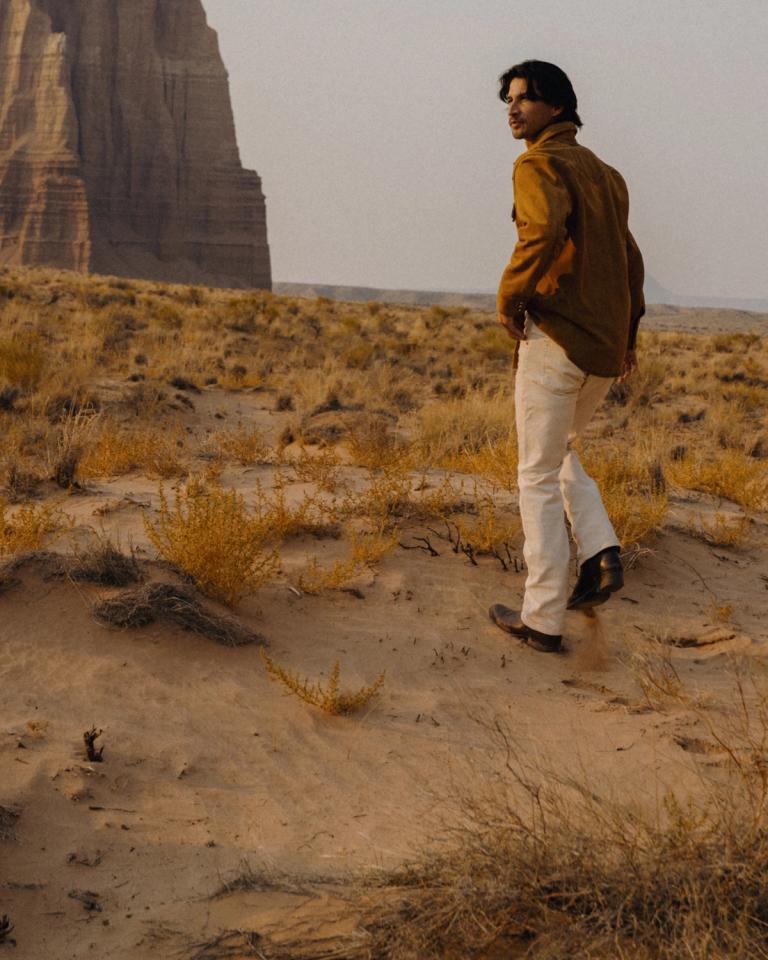 A man in a button-down shirt and light-colored trousers walks through a sandy desert landscape near a large rock formation.