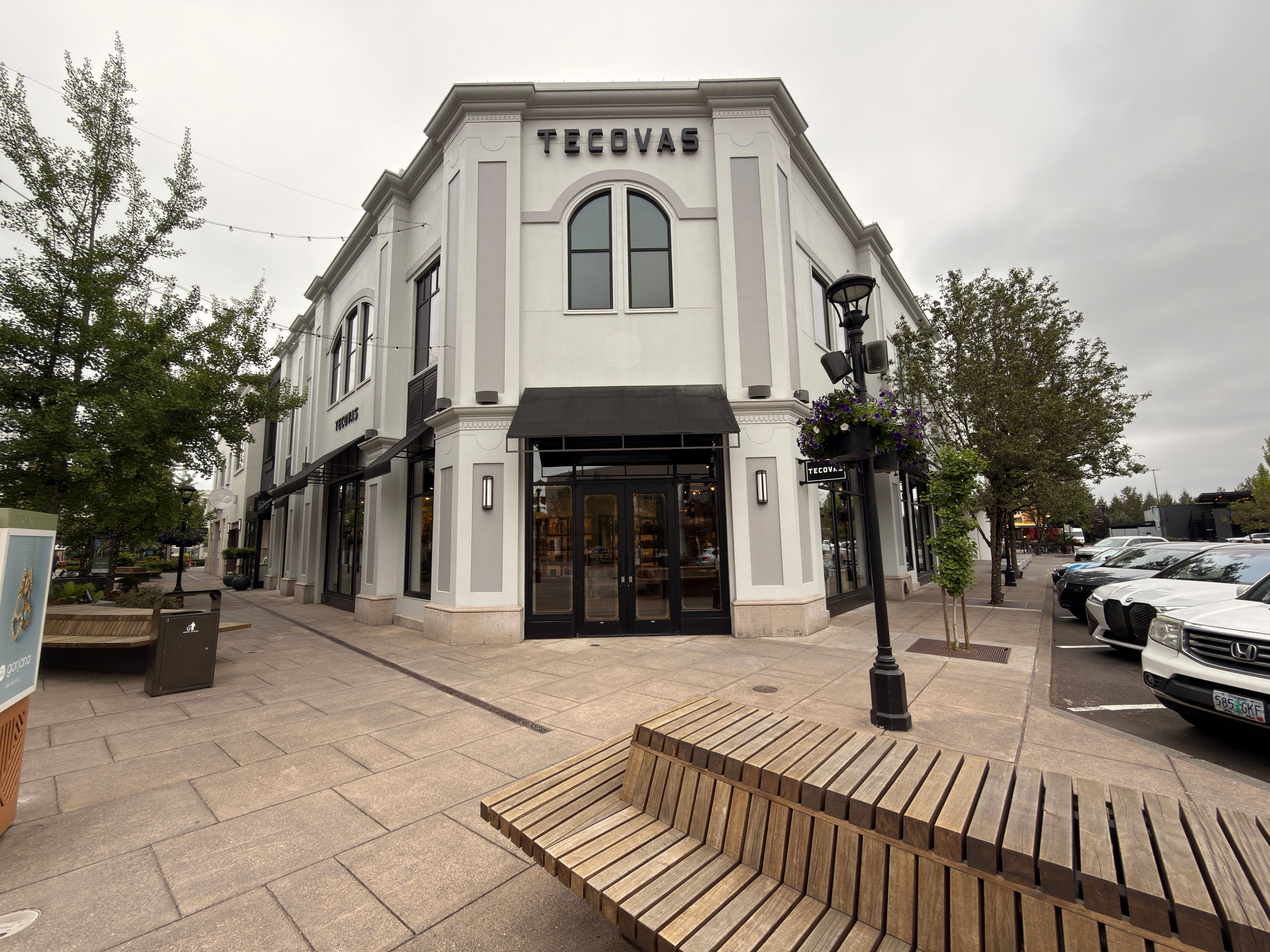 A corner view of a two-story Tecovas store with large windows, black awnings, and nearby parked cars. Benches and trees line the sidewalk under a cloudy sky.