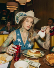 A person with tattoos and a cowboy hat pours syrup on waffles at a diner table. They hold a red drink. Others are seated in the background. Dollar bills are on the table.