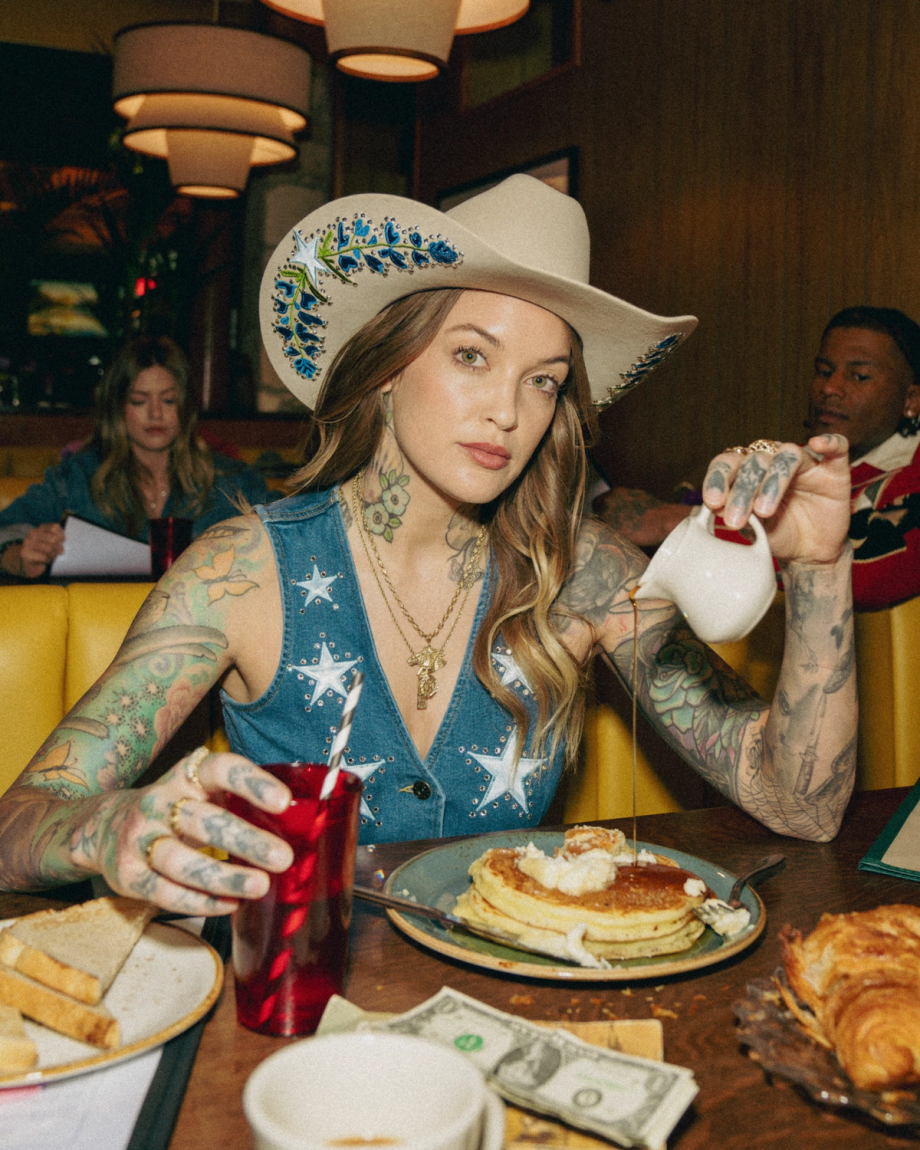 A person with tattoos and a cowboy hat pours syrup on waffles at a diner table. They hold a red drink. Others are seated in the background. Dollar bills are on the table.