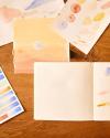 Kelly Colchin's watercolor sketches and painting of pastel blue and orange sunset laid out on a wooden tabletop.