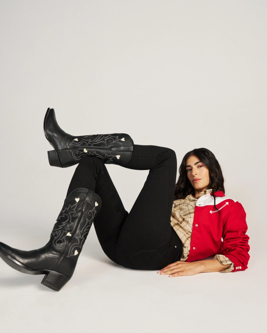 The Annie in Midnight Cowhide. A woman in a red jacket, plaid shirt, black pants, and Midnight cowhide cowboy boots lies on her back with one leg bent and the other extended against a plain studio background.