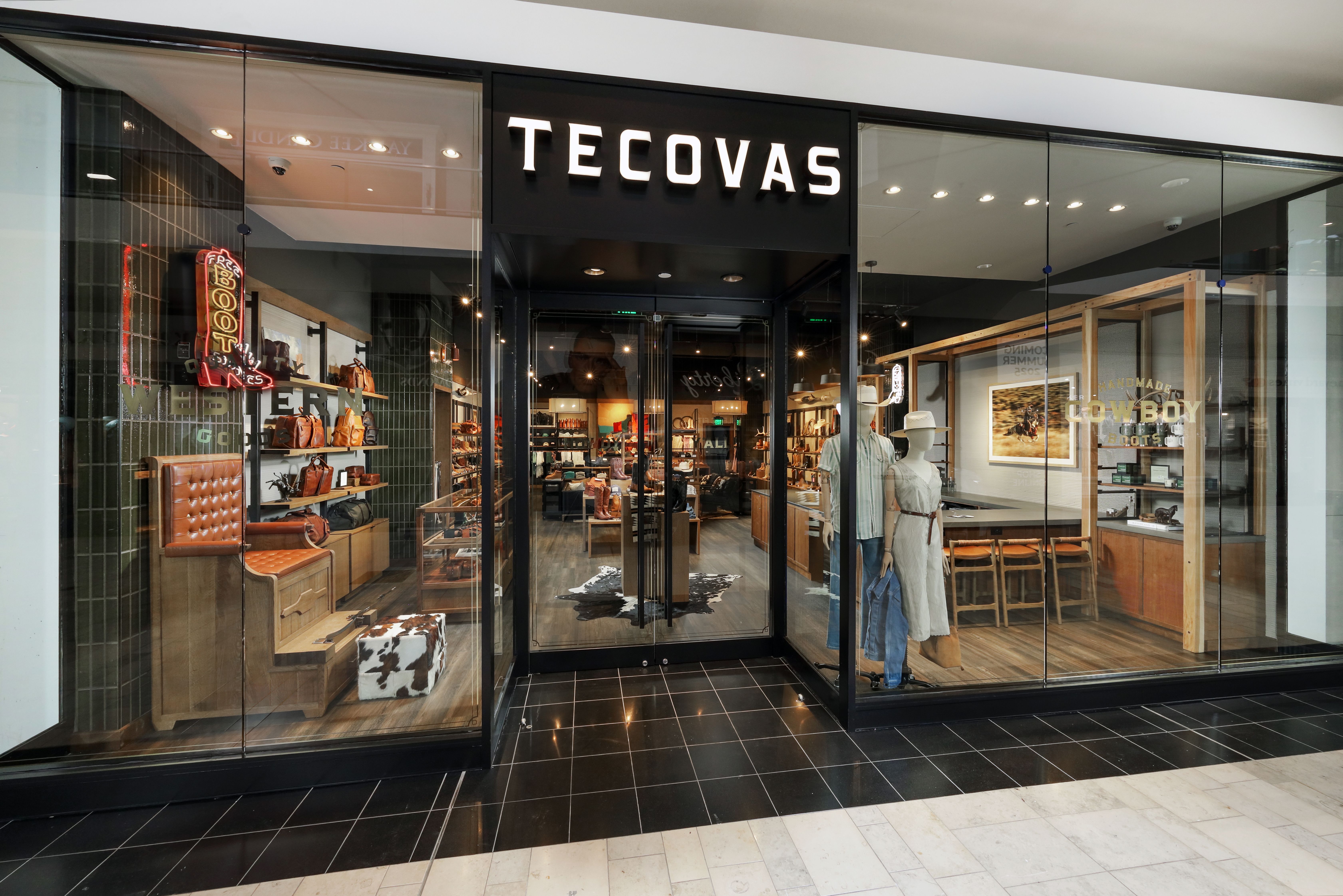 A glass storefront displays the Tecovas store, featuring boots, leather goods, and western-themed decor inside a shopping mall.