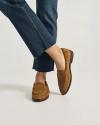 A person wearing frayed hem blue jeans and brown suede loafers stands with one foot crossed over the other against a plain background.