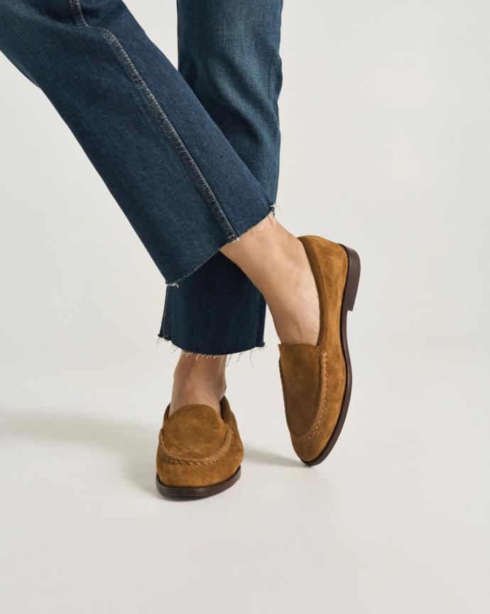 A person wearing frayed hem blue jeans and brown suede loafers stands with one foot crossed over the other against a plain background.