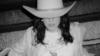 A person wearing a large cowboy hat and light-colored clothing is seen in a blurred, black-and-white photo, with their face partially obscured by the hat.