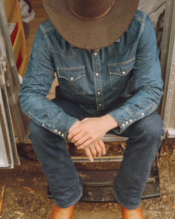 A person in denim clothing and a cowboy hat sits on a step with hands clasped, head down, and orange boots visible.