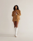 A person stands against a plain background wearing a tan fringe jacket, matching skirt, and white cowboy boots.