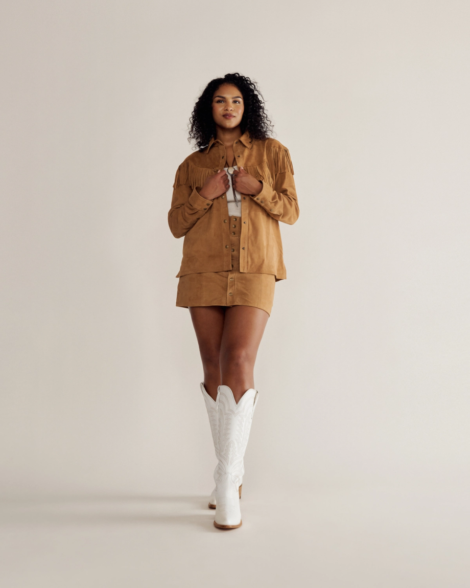 A person stands against a plain background wearing a tan fringe jacket, matching skirt, and white cowboy boots.