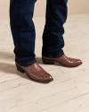 A person wearing brown ostrich leather cowboy boots and dark blue jeans standing on a light wood floor.