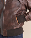 Close-up of a person wearing a brown leather jacket with their hand in the jacket pocket. The jacket has a visible zipper, reinforced stitching, and ribbed cuffs and waistband. Blue jeans are also visible.