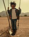 A young child stands on a swing in a desert playground, wearing a denim jacket, jeans, and cowboy boots, with mountains in the background.