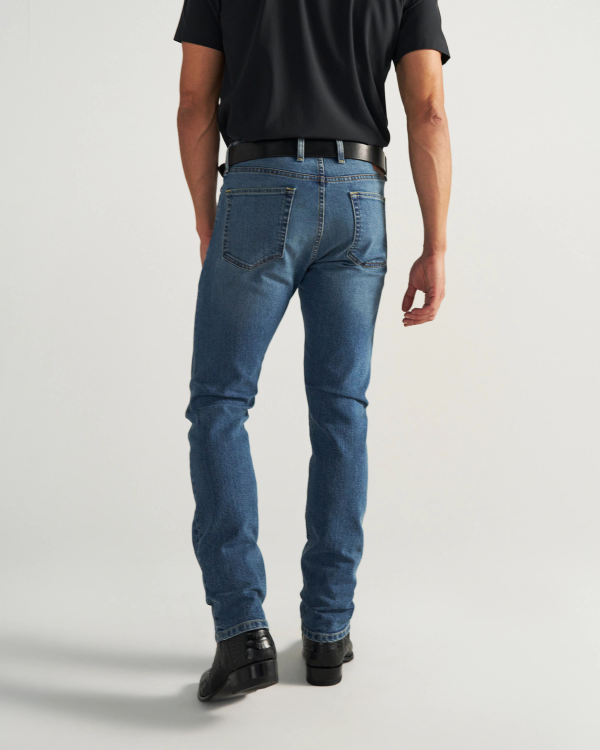 Man wearing a black shirt, blue jeans, black belt, and black shoes, standing with his back to the camera against a plain background.