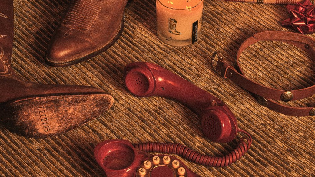 A pair of cowboy boots, a lit candle, vintage red rotary phone, dog collar, and stacked gift boxes are arranged on a striped carpet near a patterned sofa.
