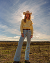 A woman in a cowboy hat and denim jeans stands in an open field, with a cloudy sky above her.