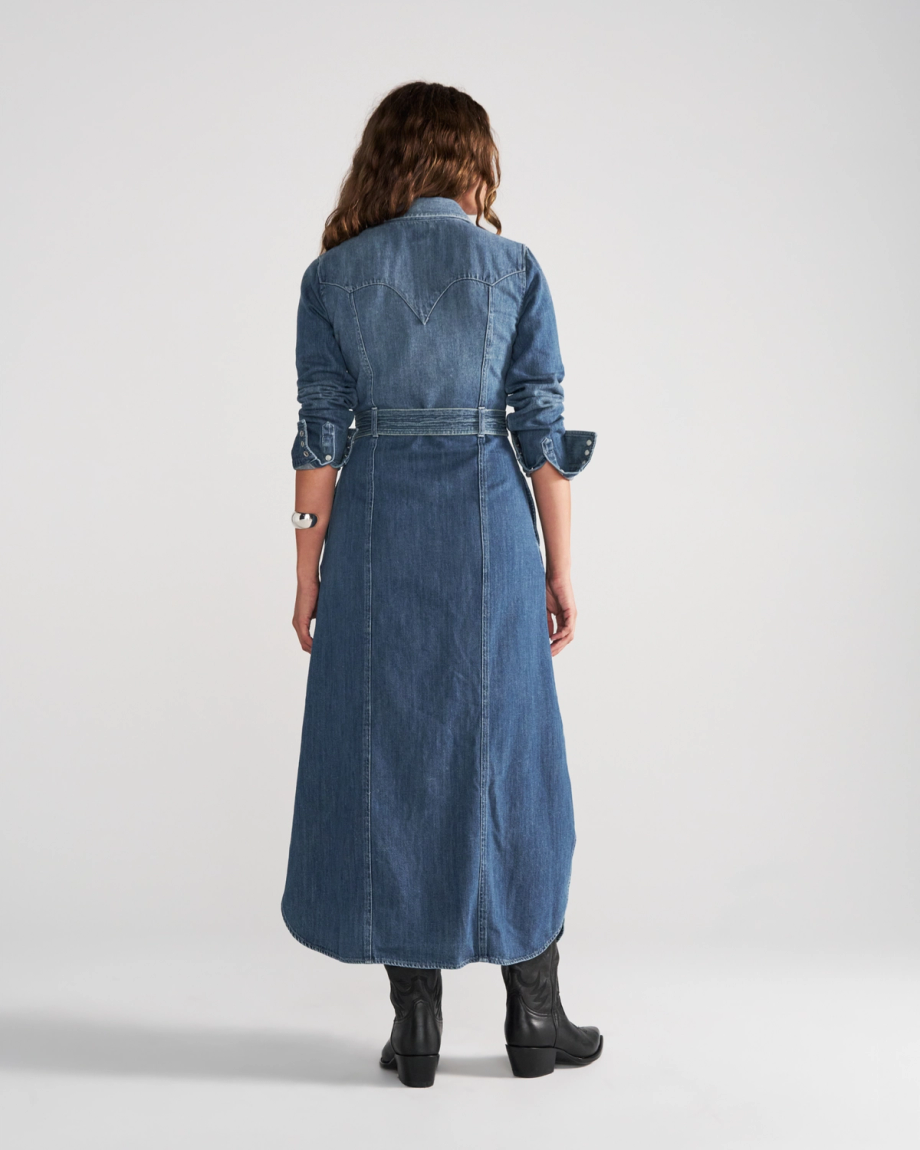 Back view of a person wearing a long-sleeved denim shirt dress with a waist belt and black leather boots.