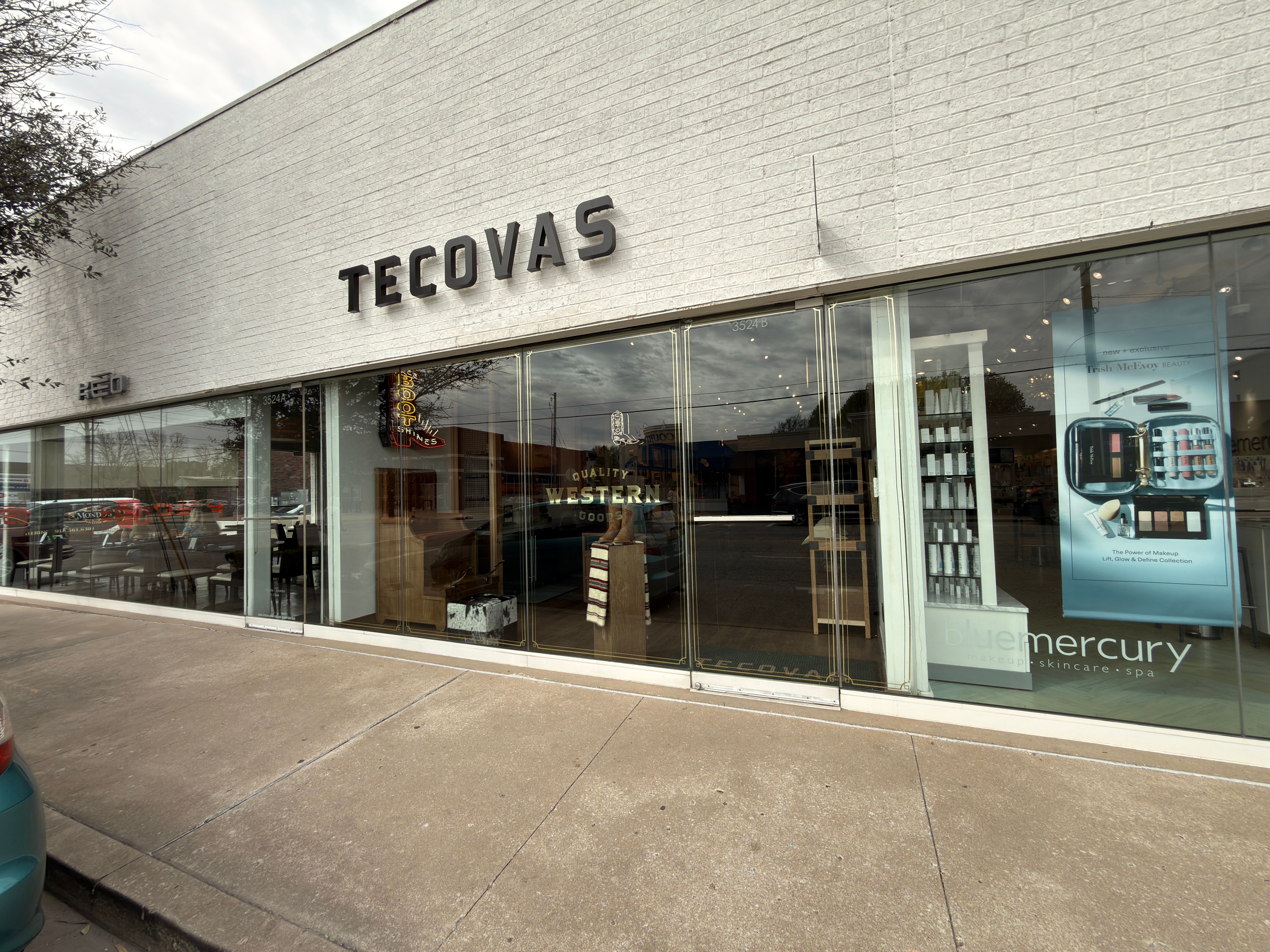 Exterior of a Tecovas store with large glass windows and adjacent Bluemercury signage; sidewalk and parked cars visible outside.