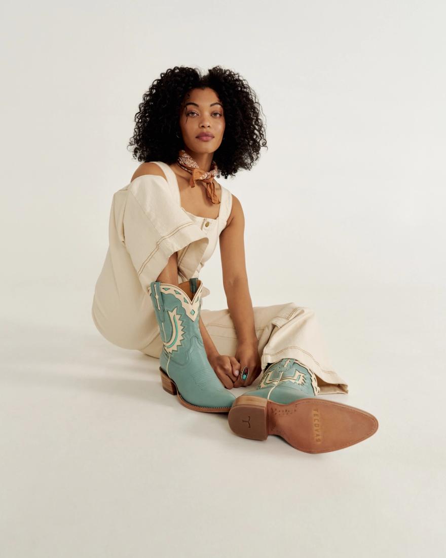 Person sitting on the floor wearing a cream outfit and turquoise cowboy boots. They're also wearing a light scarf around their neck.