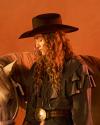 Person wearing a black wide-brimmed hat and ruffled shirt stands beside a horse against a warm-toned background.