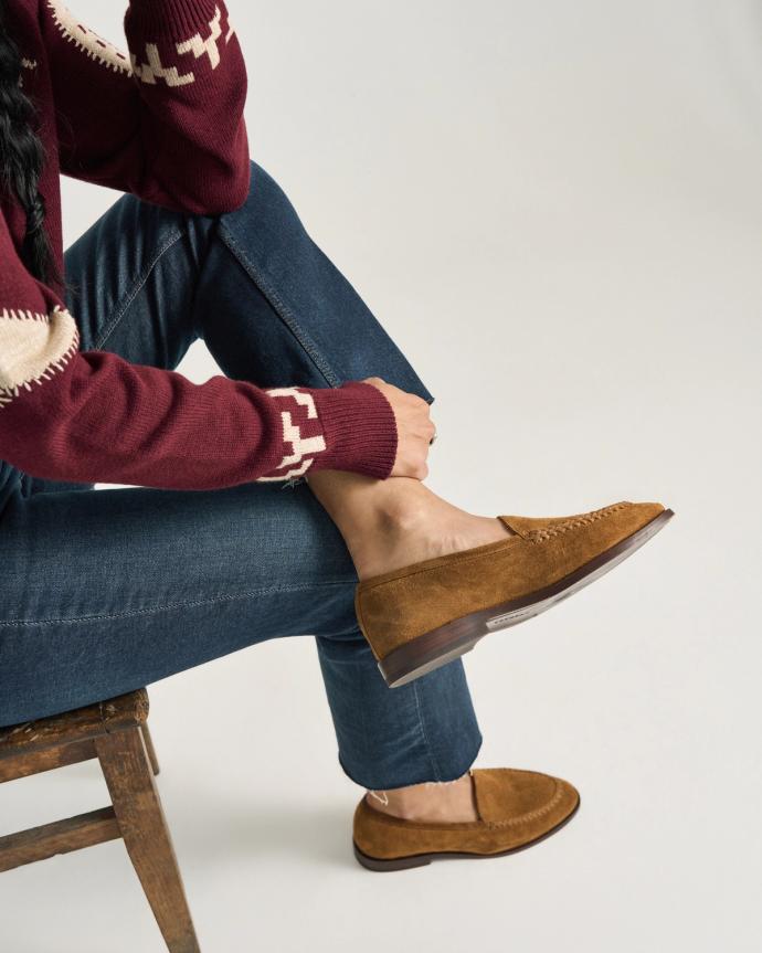Person sitting on a wooden chair with legs crossed, wearing blue jeans, a maroon sweater with a patterned sleeve, and brown suede loafers.